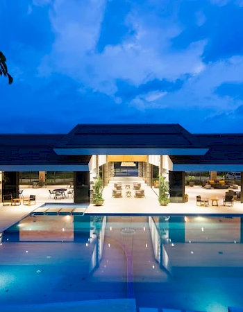 The image shows a luxurious, illuminated pool area at dusk with a modern building in the background, surrounded by clear blue skies.