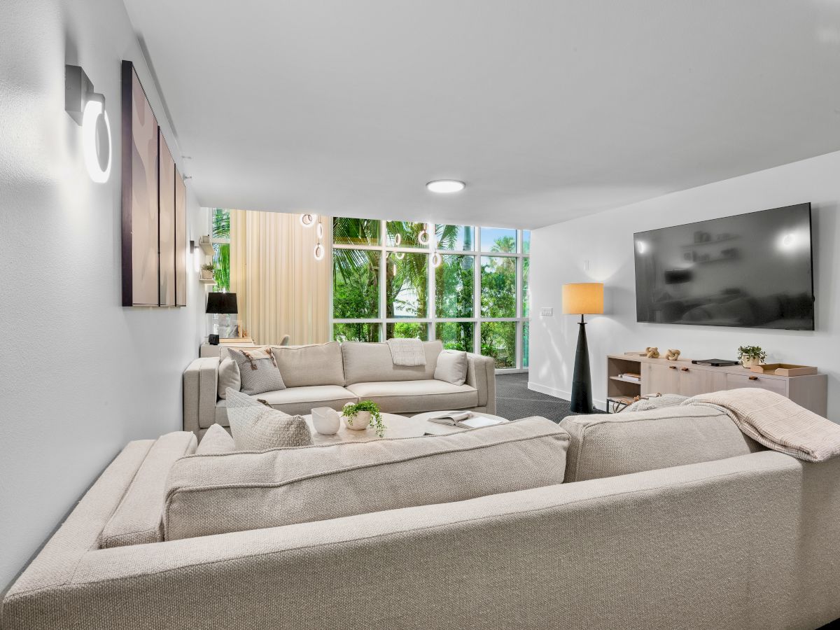 A modern living room with beige sectional sofas, a flat-screen TV, large windows, and a floor lamp, creating a cozy and inviting space.