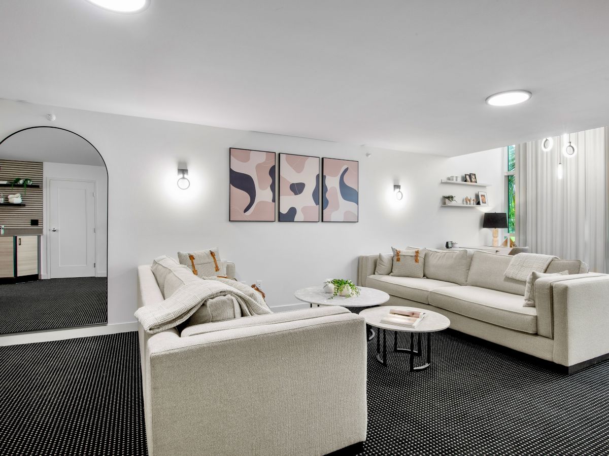 A modern living room with two beige sofas, a round mirror, abstract artwork, a small kitchenette, and large windows letting in natural light.