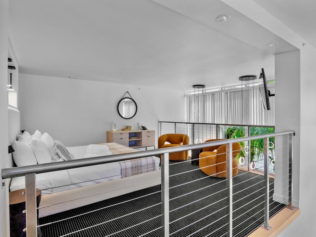 A modern loft bedroom with a bed, circular mirror, two orange chairs, and large windows with white blinds.