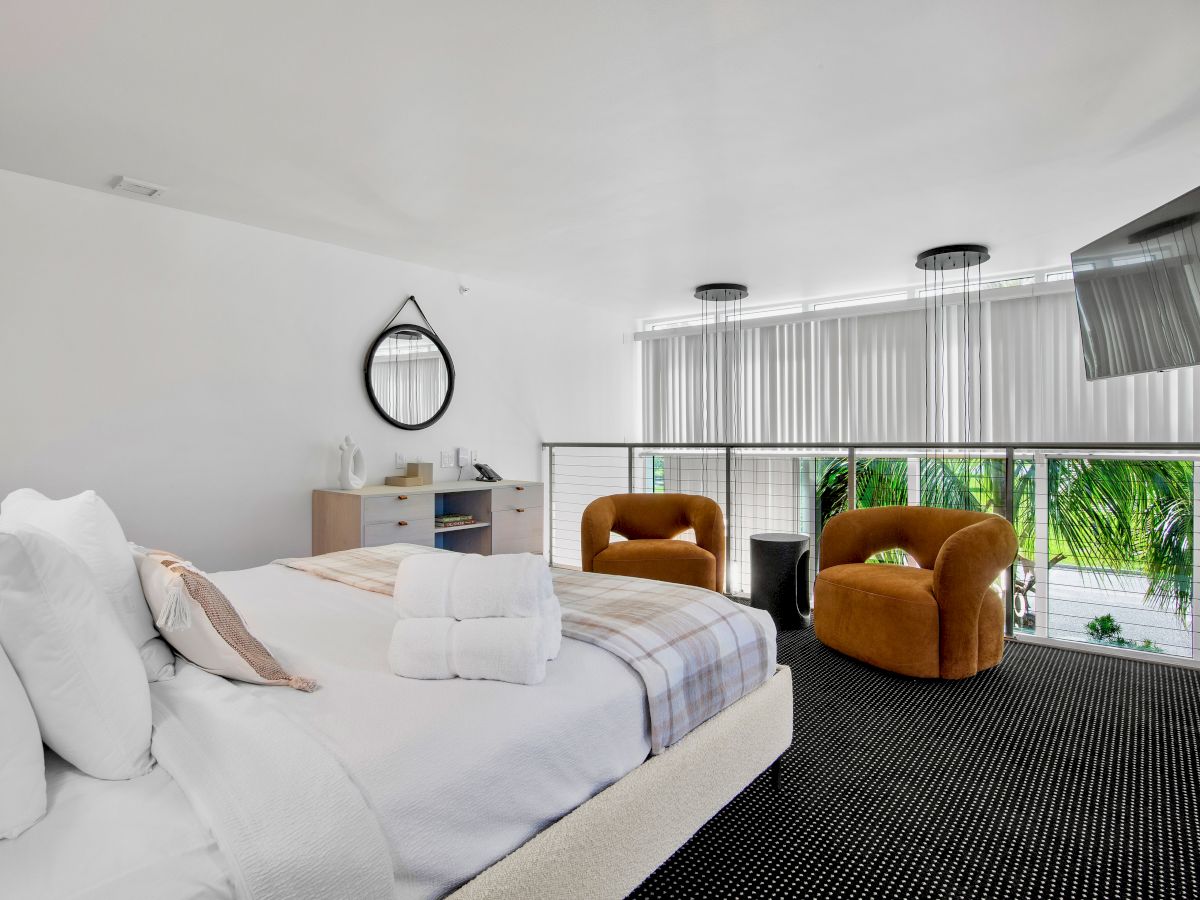 A modern bedroom with a large bed, round mirror, two orange chairs, and a wall-mounted TV, featuring large windows with blinds.