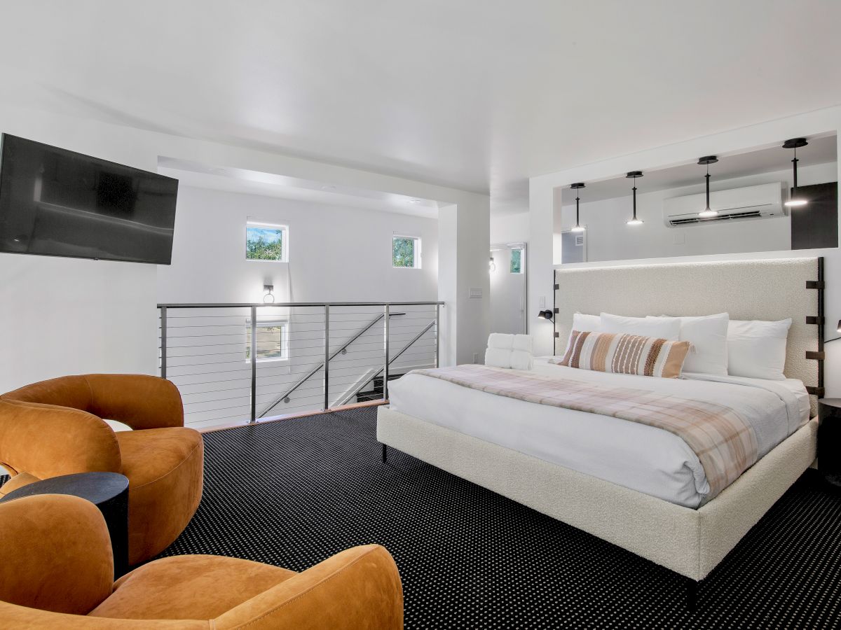 A modern bedroom with a large bed, two orange chairs, a wall-mounted TV, and minimalist decor in a bright, neutral-toned room.