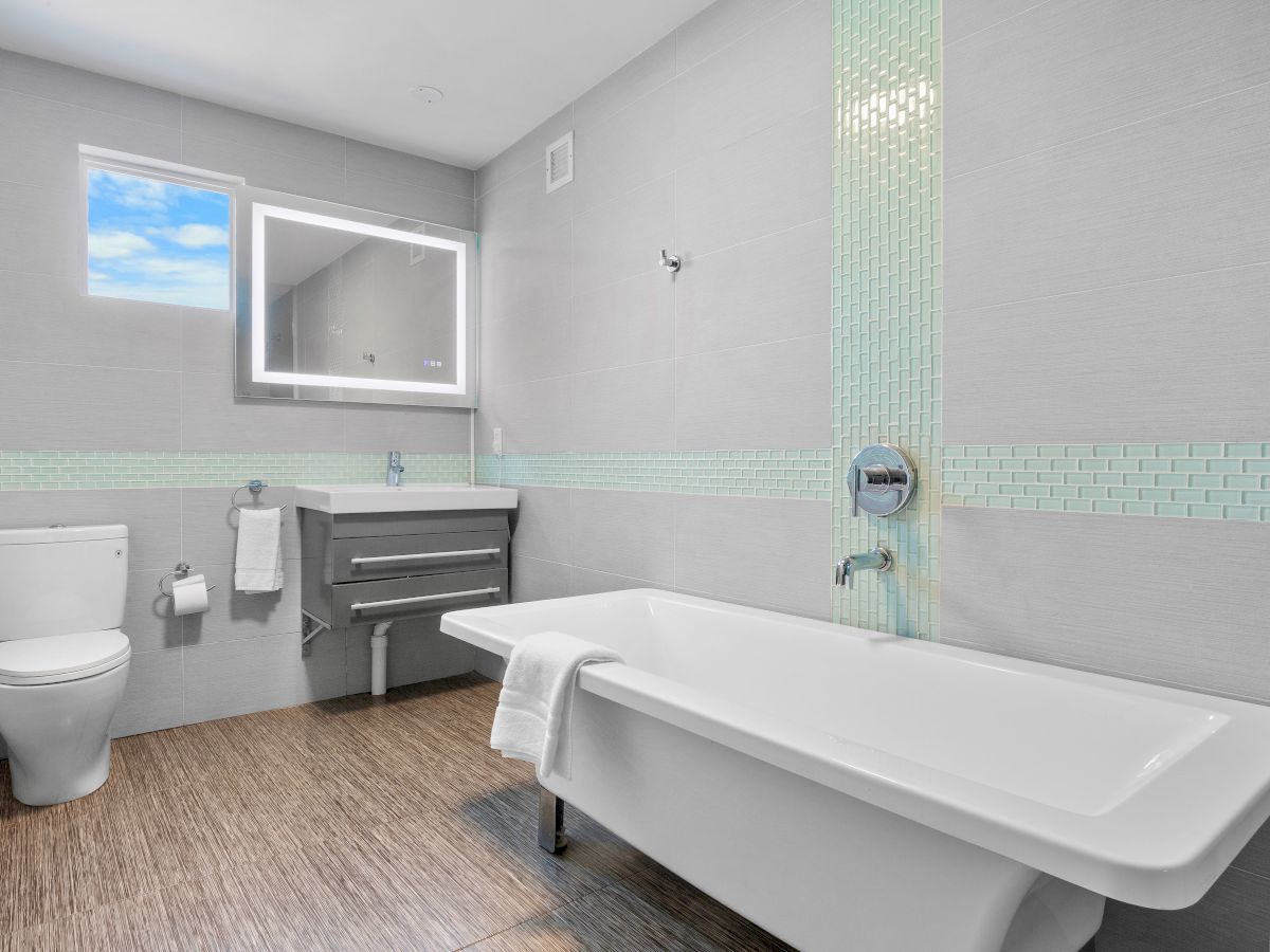 A modern bathroom with a bathtub, toilet, and sink, featuring gray tiles and a window showing a blue sky.