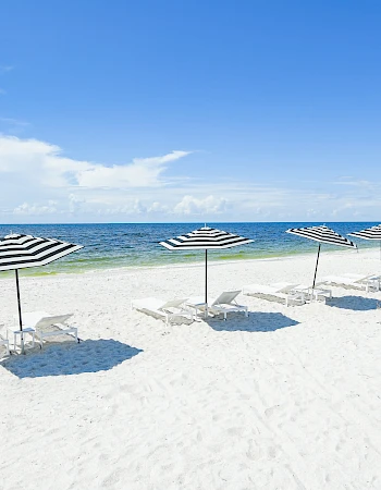 Row of striped beach umbrellas with empty chairs on a white sandy beach, calm blue sea, and clear sky in the background.