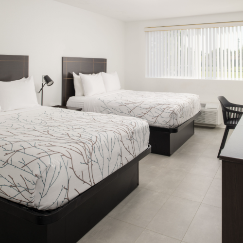 The image shows a hotel room with two double beds, a desk with a chair, a lamp, and a TV on a dresser, under a large window with blinds.