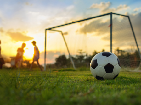 A soccer ball lies on grassy field with a goal in the background. Three figures are silhouetted against a vibrant sunset sky.
