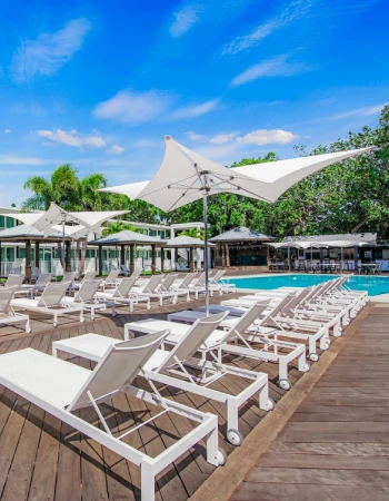 A poolside area with white lounge chairs, umbrellas, and a wooden deck surrounded by greenery and a clear blue sky.