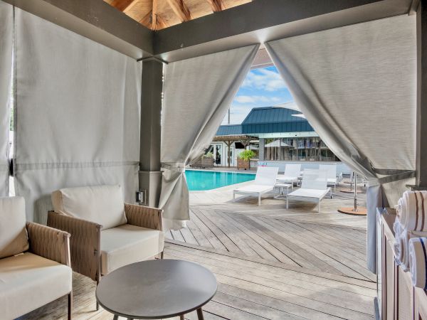 A cozy poolside cabana with comfortable seating and curtains, overlooking a pool and lounge chairs under a blue sky.